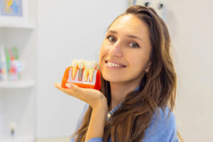 Smiling young woman holding a dental implant model, demonstrating tooth replacement options and implant-supported restoration in a dental office.